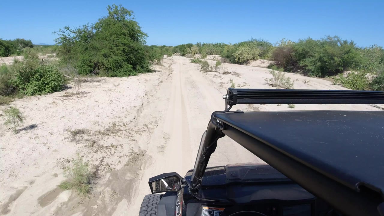 The Jeep remains in the lower right foreground as the sandy Los Cabos track narrows between pale banks and scattered shrubs under clear blue sky, with no visible people.