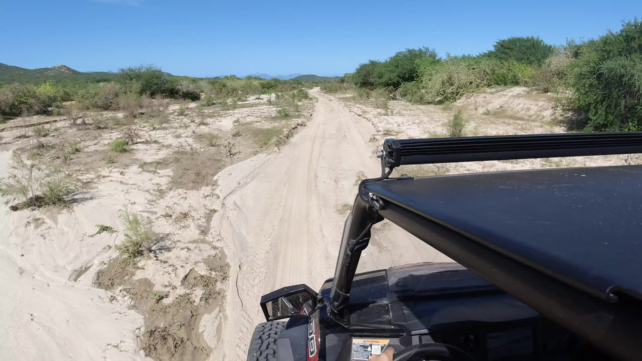 Forward view from a Wrangler on a sandy Los Cabos, Mexico track, with dry scrub on both sides, low hills ahead, bright blue sky, and no visible people.