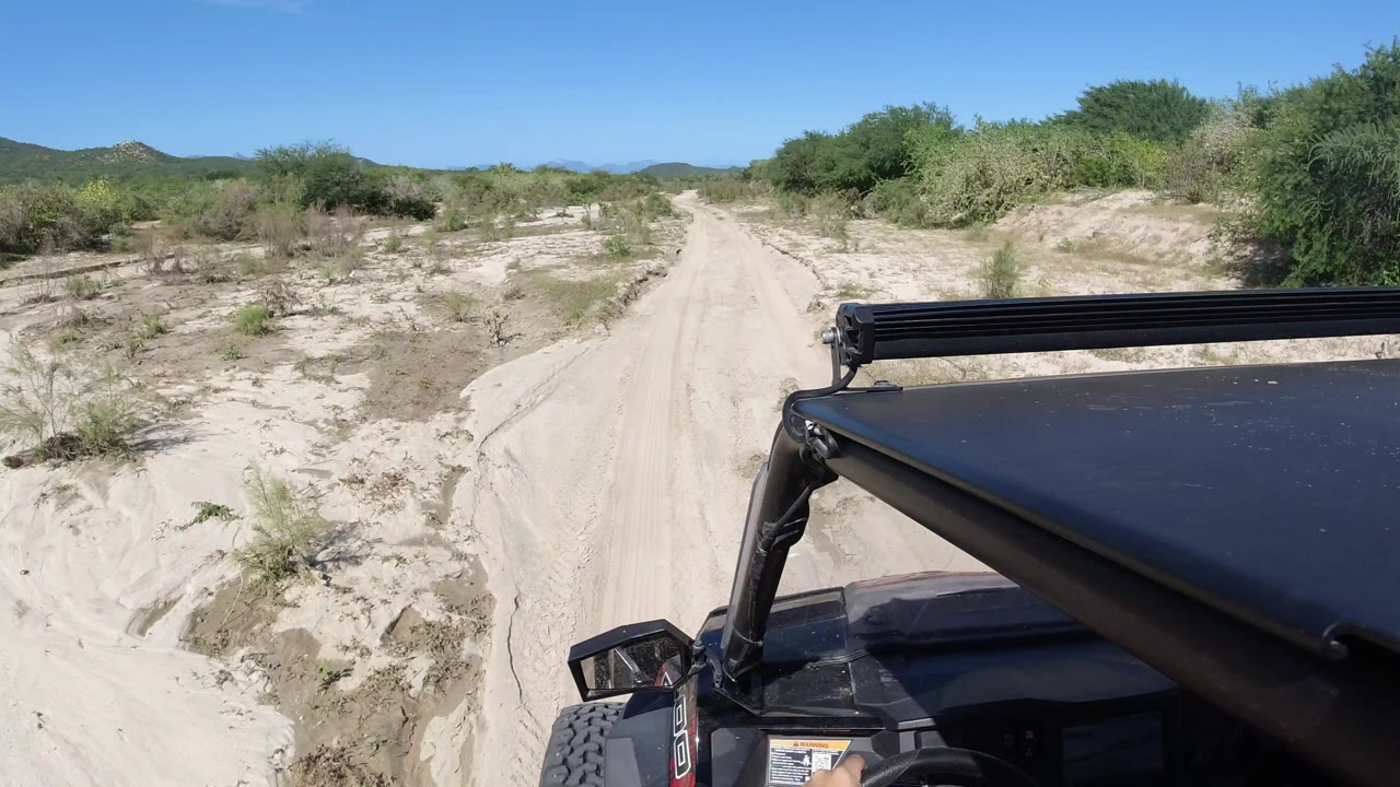 The Wrangler continues along the pale sandy road in Los Cabos, Mexico, with the black hood at right, sparse dry plants along the track, distant hills, and no visible people.