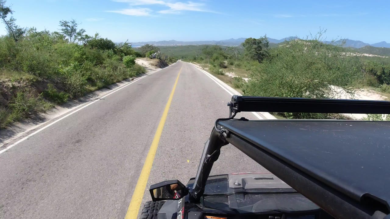 Straight paved road in Los Cabos, Mexico, bordered by dense green shrubs, with the vehicle roof edge and side mirror fixed in the lower foreground.