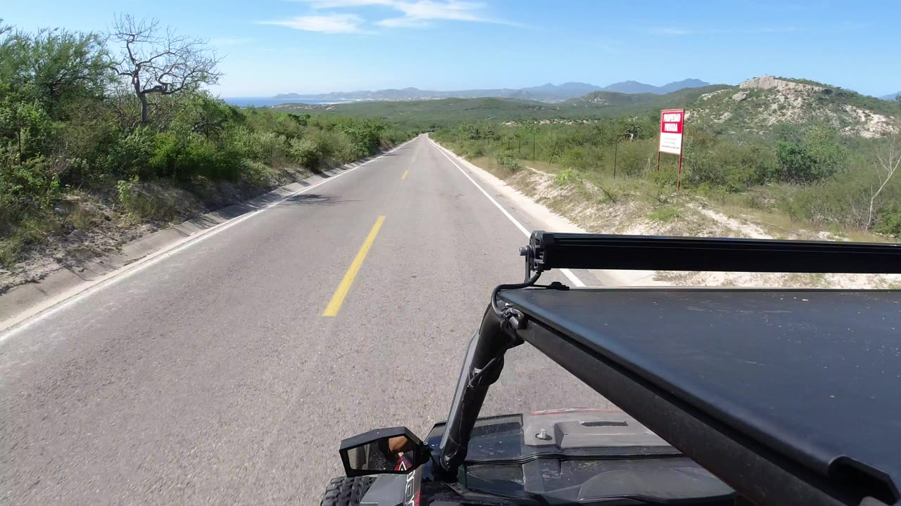 Opening view down a straight paved road in Los Cabos, Mexico, with green brush on both sides, distant hills, a roadside sign, black vehicle hardware in the foreground, and no visible people.