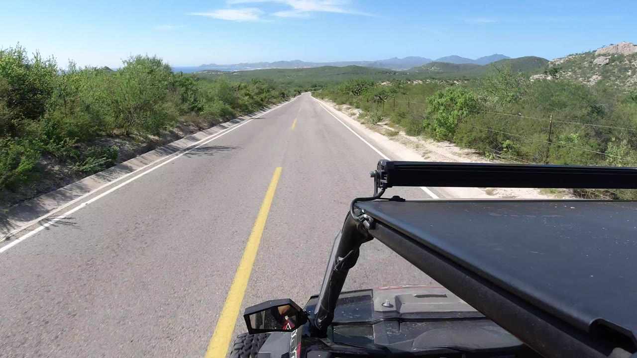 Forward view over vehicle hardware on a paved Los Cabos road, with desert bushes, pale shoulders, distant hills, blue sky, and no visible people.