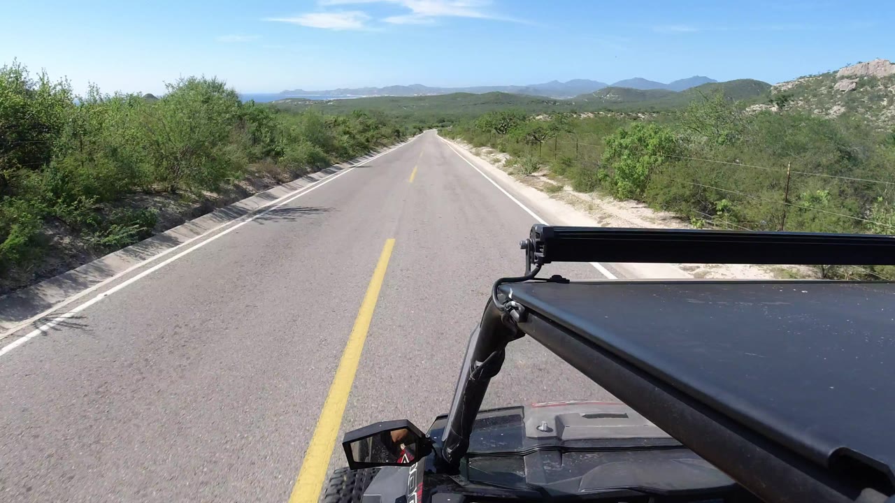 Straight Los Cabos paved road receding toward low hills, bordered by dense green brush and sandy roadside, with vehicle hardware at lower right and no visible people.