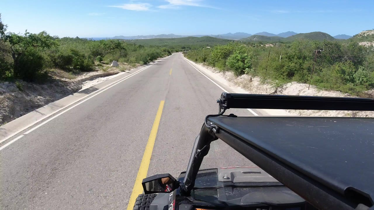 Start of the Los Cabos road view, with the black Jeep roof and roll bar in the foreground, a yellow center line leading downhill, green brush on both sides, distant hills, blue sky, and no visible people.