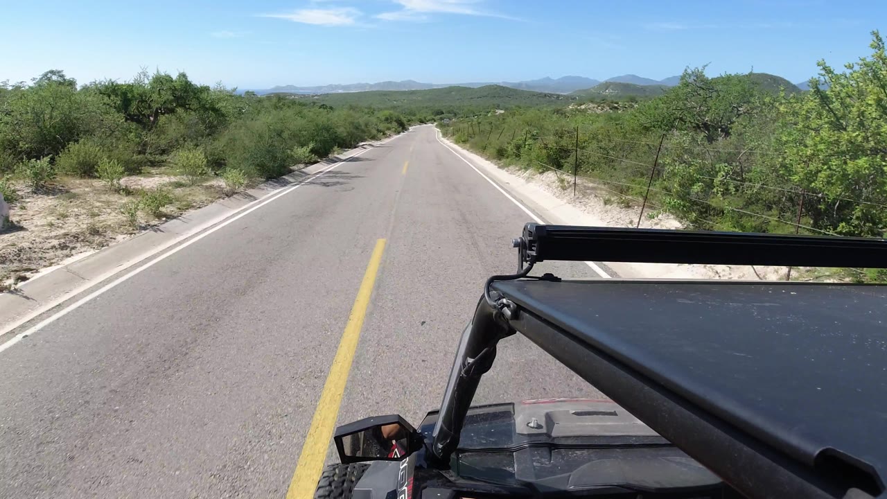 Vehicle-mounted view of an empty road curving past brush and low mountains.
