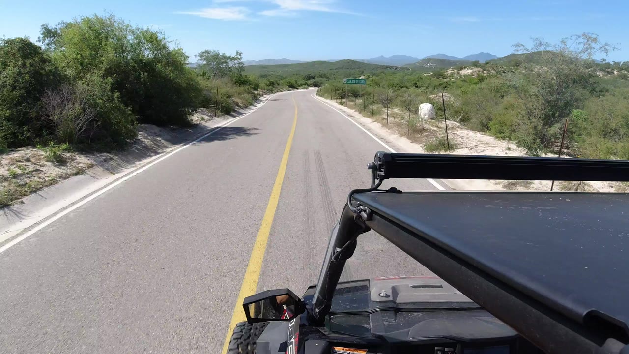 Forward road view from a vehicle with scrub and low hills ahead.