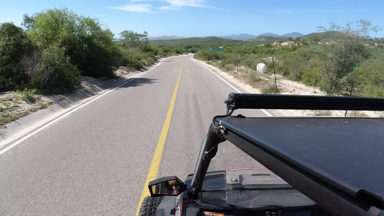 Los Cabos driving view one second in, with the road stretching ahead between sandy shoulders, dense shrubs, distant hills, and no visible people.