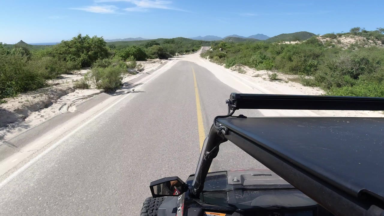 The Los Cabos road continues straight ahead under bright sun, with the black vehicle roof frame at lower right, sandy shoulders, shrubs, and no visible people.