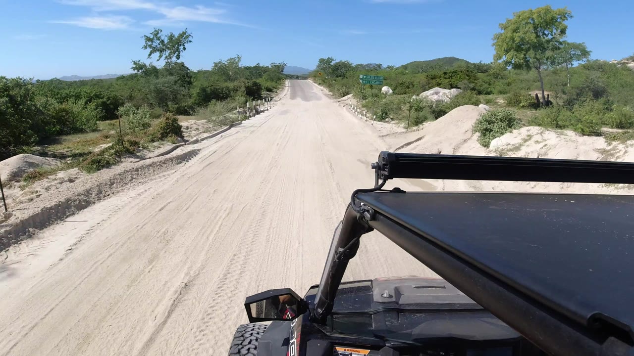 A forward Jeep-mounted view in Los Cabos, Mexico, shows the sandy road continuing between green roadside brush, with a small roadside sign area visible ahead and no visible people.