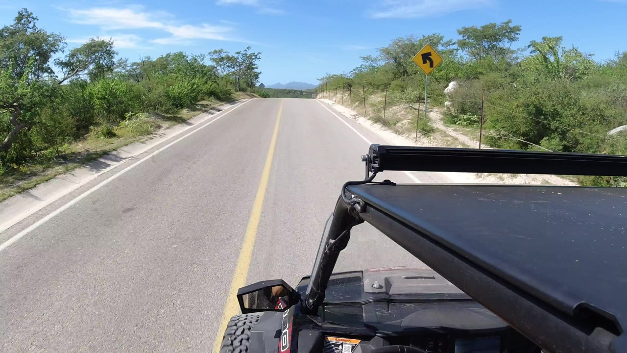 Forward view from the Wrangler on a sunlit Los Cabos road, with a yellow centerline stretching ahead, scrubby green vegetation on both sides, the vehicle hood in frame, and no visible people.