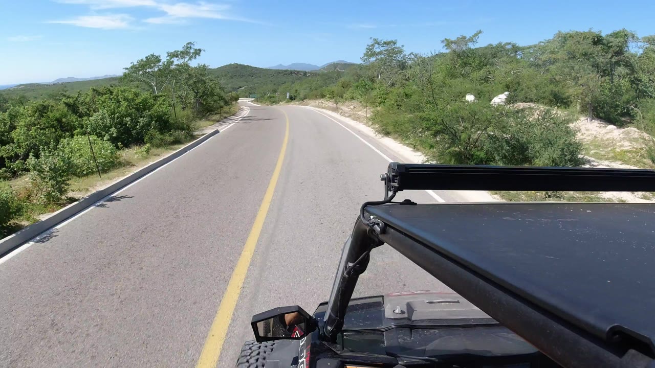 The RZR continues along the Los Cabos road, with the vehicle roof edge and mirror in the lower frame, scrub vegetation close to the shoulders, distant hills, and no visible people.