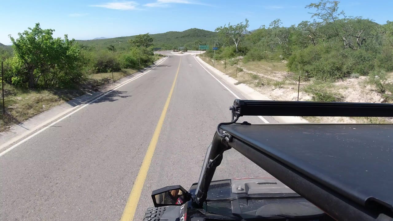 Vehicle POV on a narrow road with green roadside brush and low hills ahead.