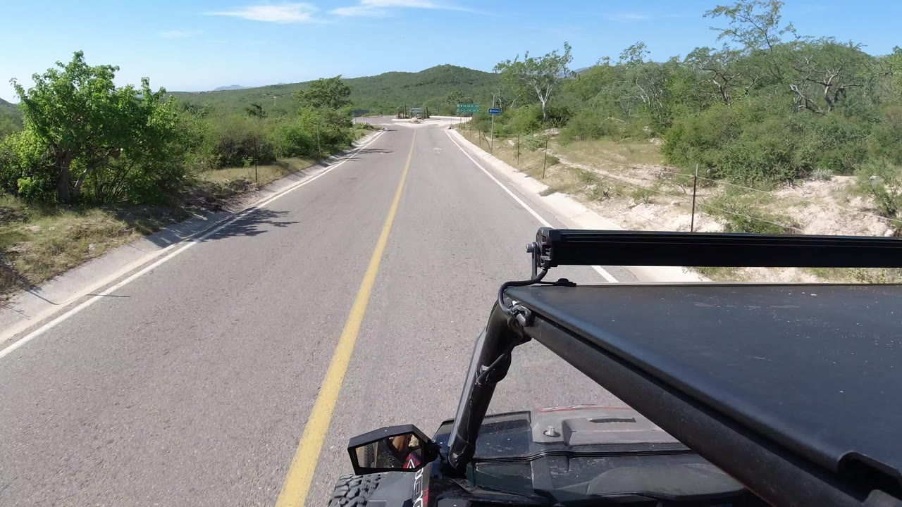 Los Cabos, Mexico road POV with the vehicle roof at lower right, a yellow centerline leading forward, scrubby vegetation on both sides, and no visible people.