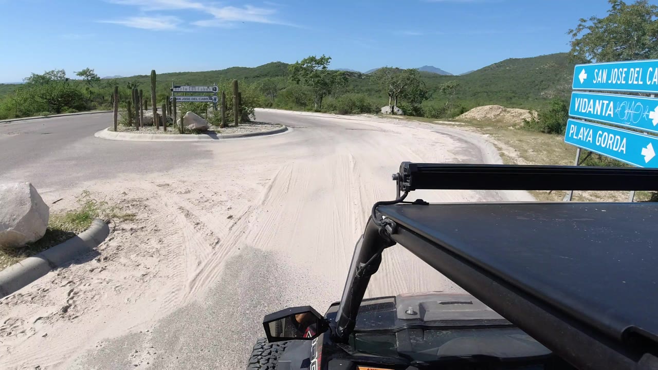 Mounted Jeep perspective in Los Cabos, Mexico, with blue signs close to the vehicle, a pale paved road curving right, desert planting, and no visible people.