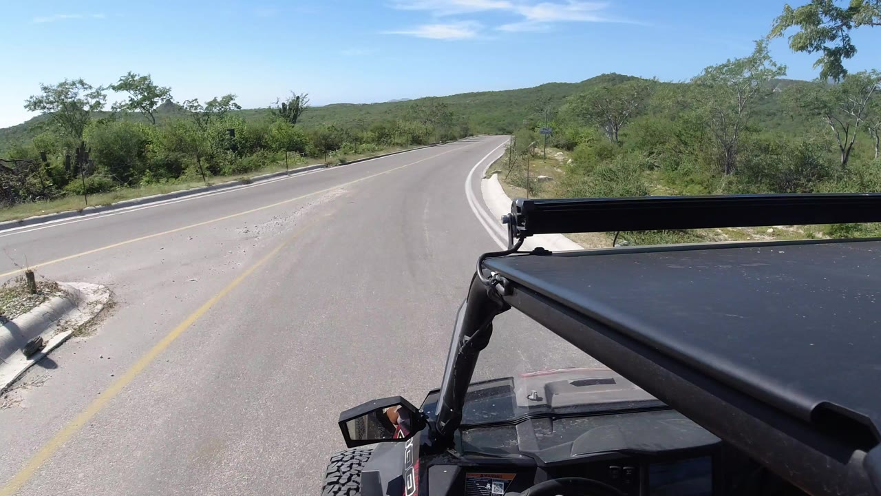 Forward view from a Jeep on a curving Los Cabos highway, with the roll bar and roof in the foreground, green brush on both sides, low hills beyond, and no visible people.