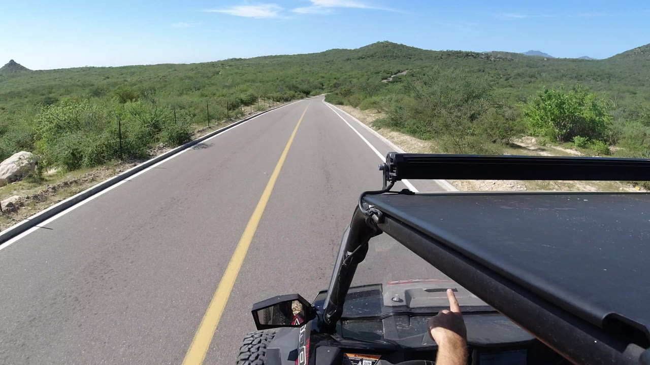 Vehicle POV on an empty road through green hills with a pointing hand in foreground.