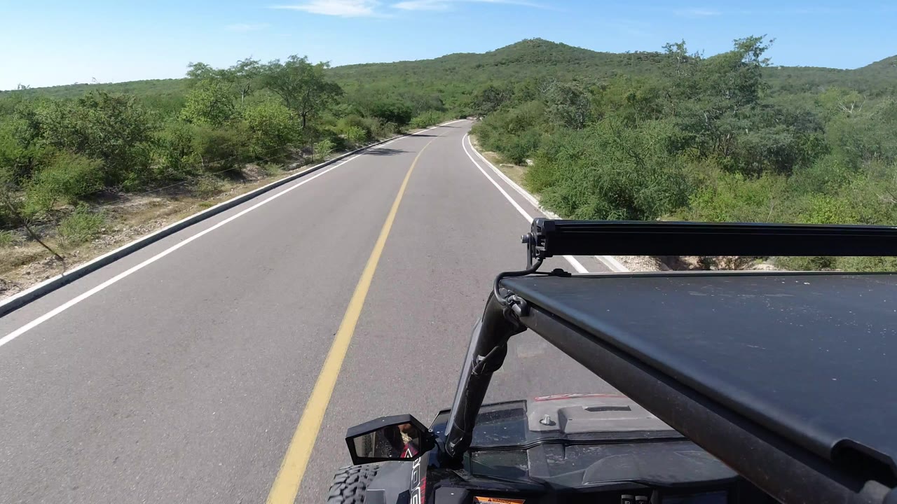 Forward view from a vehicle on a paved Los Cabos road, with a yellow center line, green roadside bushes, low hills ahead, and no visible people.