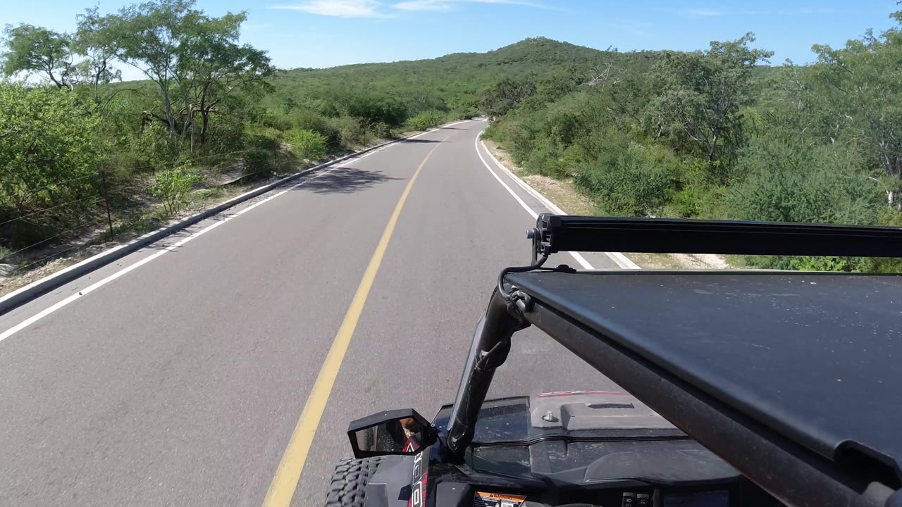 Curving paved road with bushes, hills, and vehicle rack at lower right.