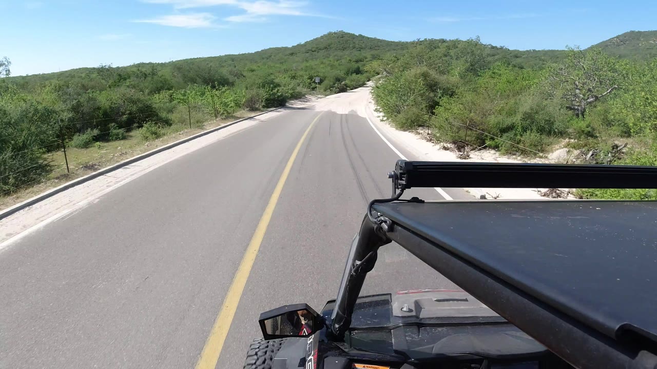 A centered Los Cabos road view shows the yellow line running forward between sandy shoulders, scrub trees, distant hills, and the black vehicle frame in the foreground, with no visible people.