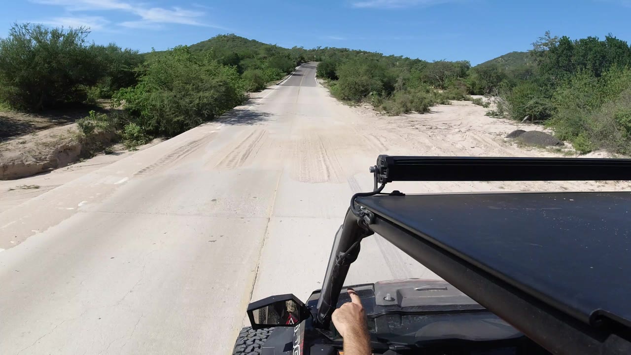 Forward view from a black Jeep on a pale dusty road in Los Cabos, Mexico, with green roadside brush, low hills, blue sky, and no visible faces.