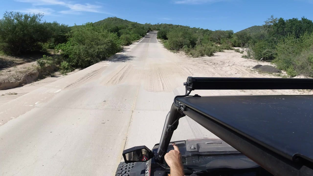The Jeep continues along the light dirt road in Los Cabos, Mexico, with dense green brush on both sides, distant hills, clear sky, and no visible faces.