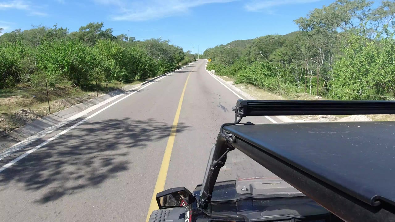 Forward view from a Jeep on a two-way road in Los Cabos, Mexico, with green trees along both shoulders, low hills ahead, bright blue sky, and no visible people.
