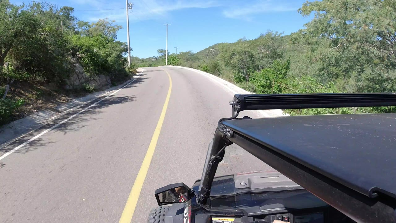 The Jeep roof and light bar fill the lower foreground as the road runs ahead through sunlit green vegetation and low hills in Los Cabos, Mexico, with no visible people.