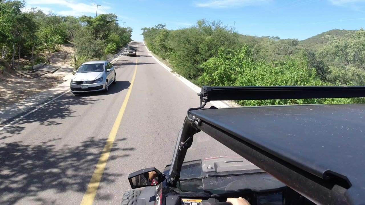 Forward-facing Wrangler view in Los Cabos, Mexico, with a white car closer to the camera, a straight paved road ahead, roadside trees, and no identifiable people visible.