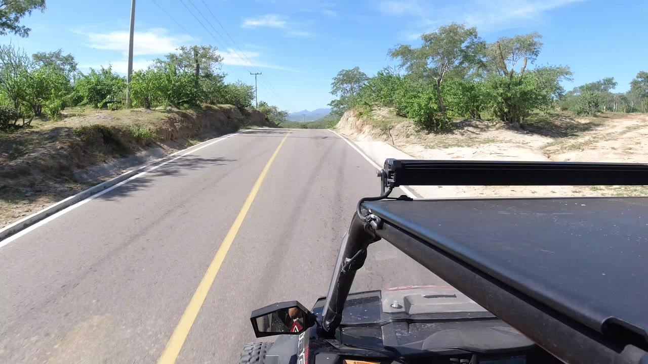A straight stretch of road in Los Cabos, Mexico runs between green vegetation and sandy banks, with the black vehicle frame visible at the lower right and no visible people.