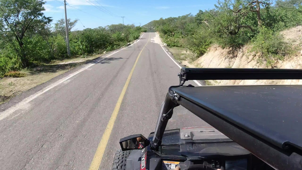 Forward view from an open vehicle on a quiet paved road bordered by green brush.