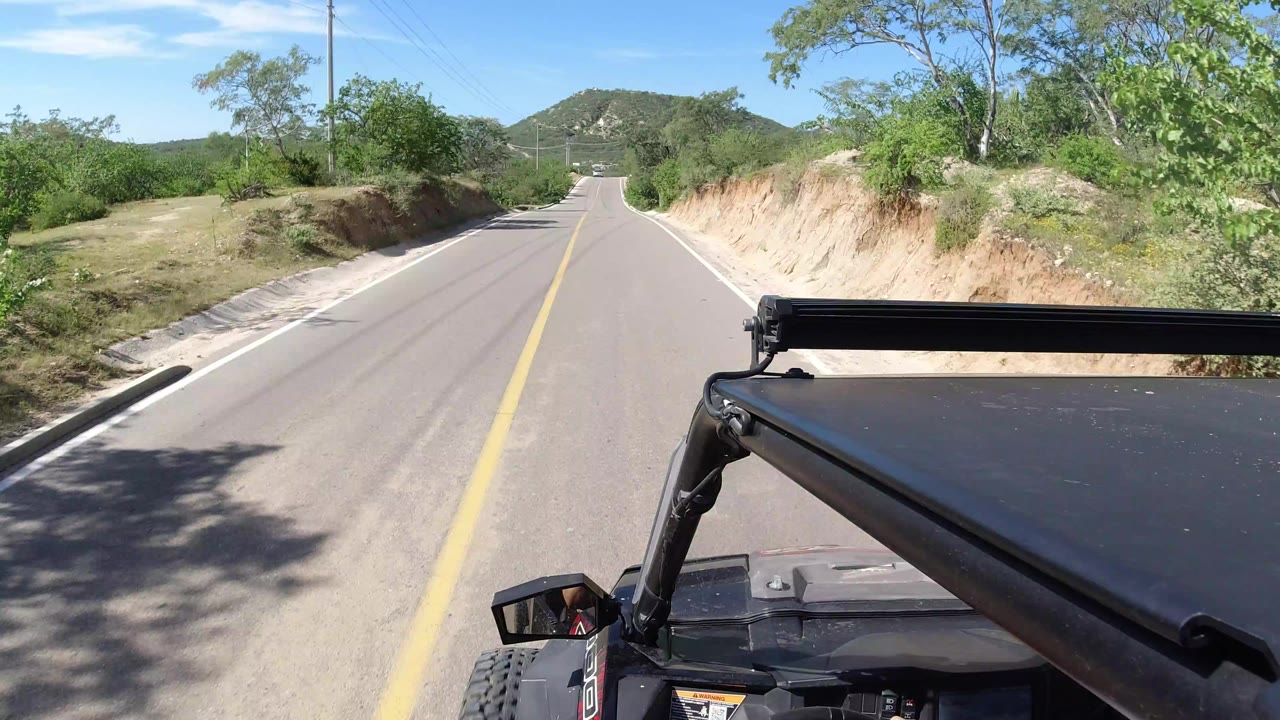 Forward-facing view from inside a Wrangler driving on a paved rural road in Los Cabos, Mexico, with a yellow center line, green roadside trees, sandy banks, low hills, and no visible people.