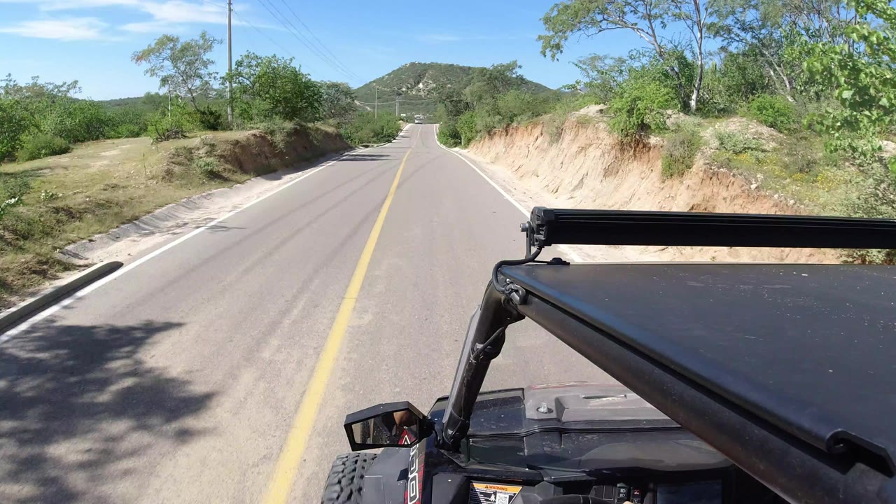 Inside-Wrangler road view in Los Cabos, Mexico, with the black roof edge and support bar filling the lower right, a narrow paved lane ahead, scrubby vegetation, sandy roadside cuts, and no visible people.