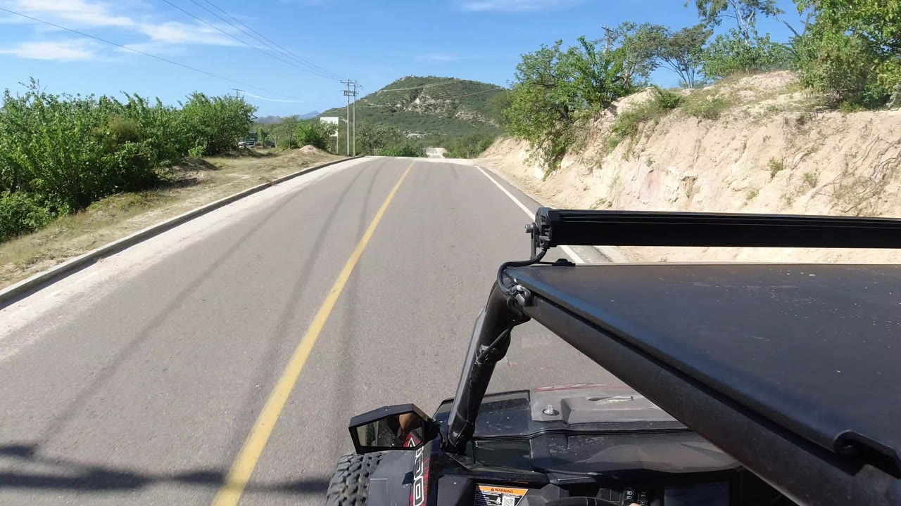 A roof-mounted Jeep perspective in Los Cabos shows the road continuing ahead beside a pale sandy embankment, green shrubs, utility wires, distant hills, and no visible people.
