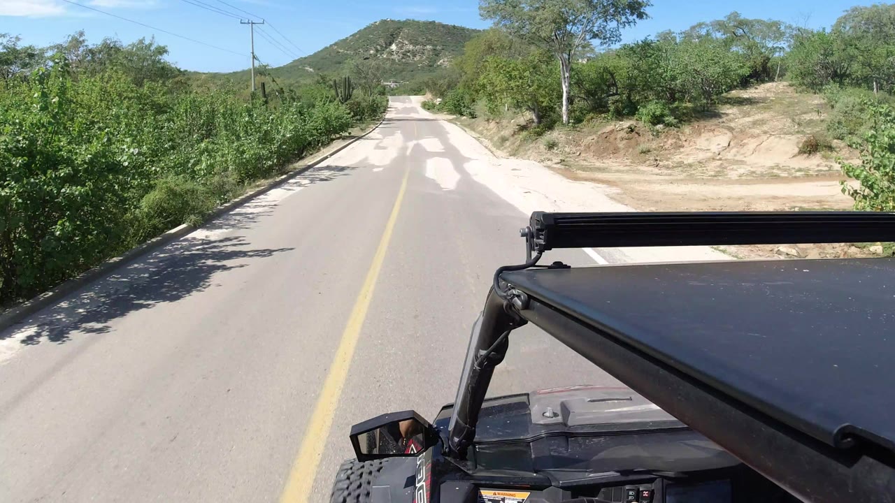A high-mounted jeep view in Los Cabos, Mexico, follows the center of the road toward a low hill, with dense green shrubs and no visible people.