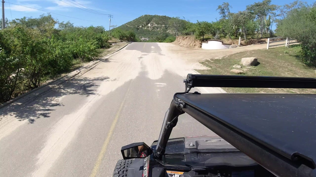 Forward-facing vehicle view on a paved Los Cabos road, with green roadside brush, a low hill in the distance, black vehicle hardware in the foreground, and no visible people.