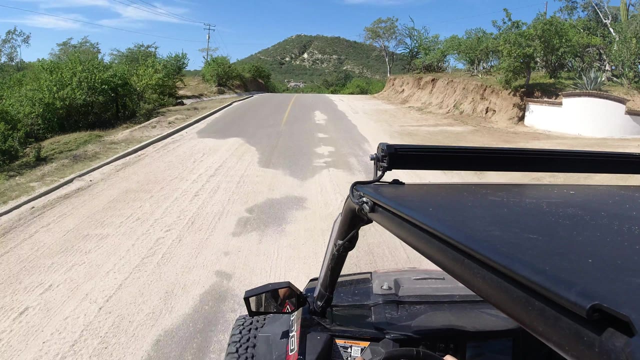 A closer forward view shows the Los Cabos road curving slightly, a tan dirt bank on the right, brush and trees along the shoulders, black vehicle hardware in front, and no visible people.