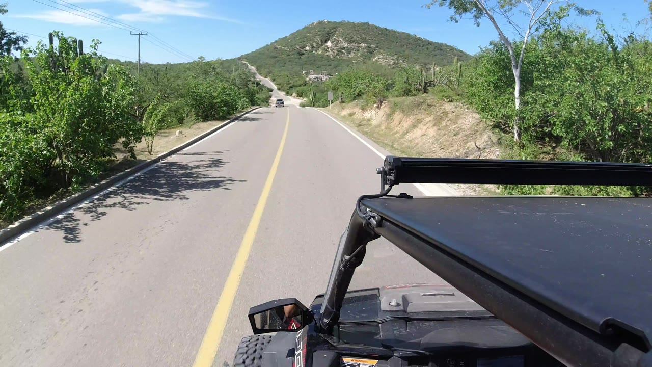A Los Cabos road continues straight ahead past dense green vegetation, with the Wrangler roof edge in the foreground and no visible people.
