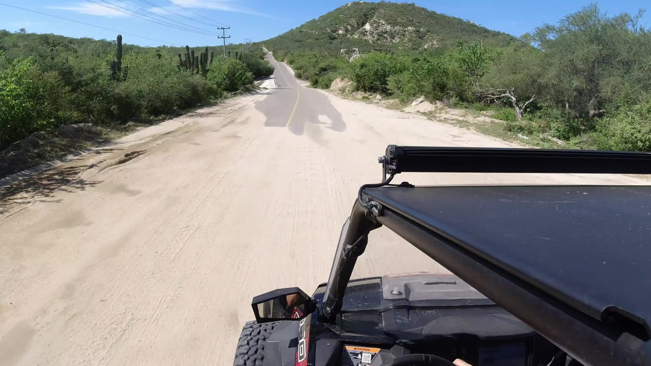 Forward view from an open vehicle on a sandy roadside beside a paved road.