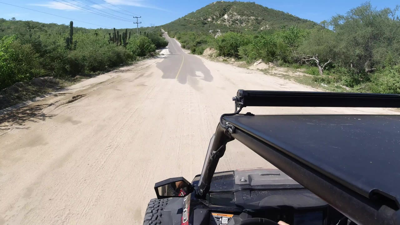 In Los Cabos, Mexico, the view looks ahead past the vehicle frame toward a narrow road bordered by sandy shoulders, dense green desert vegetation, cacti, and hillside terrain.