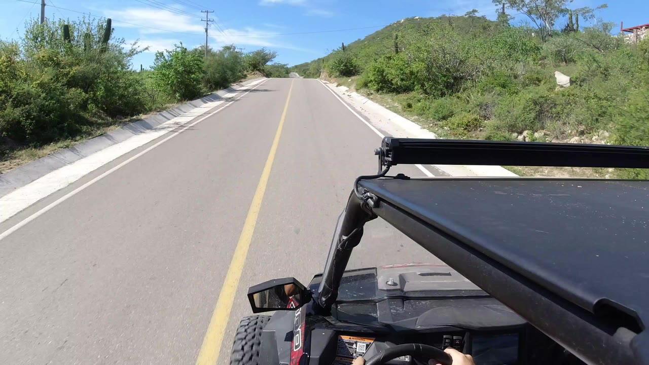 Forward-facing view from an open vehicle on a paved Los Cabos road, with a yellow center line, sandy shoulders, green brush, low hills, and no visible faces.