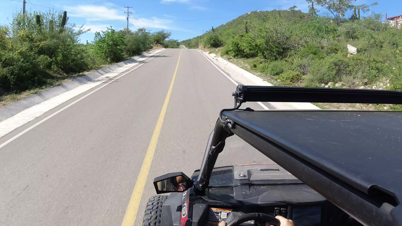 The vehicle continues along the Los Cabos road, with the black roof bar across the lower frame, dense green roadside brush, sandy drainage edges, and no visible faces.