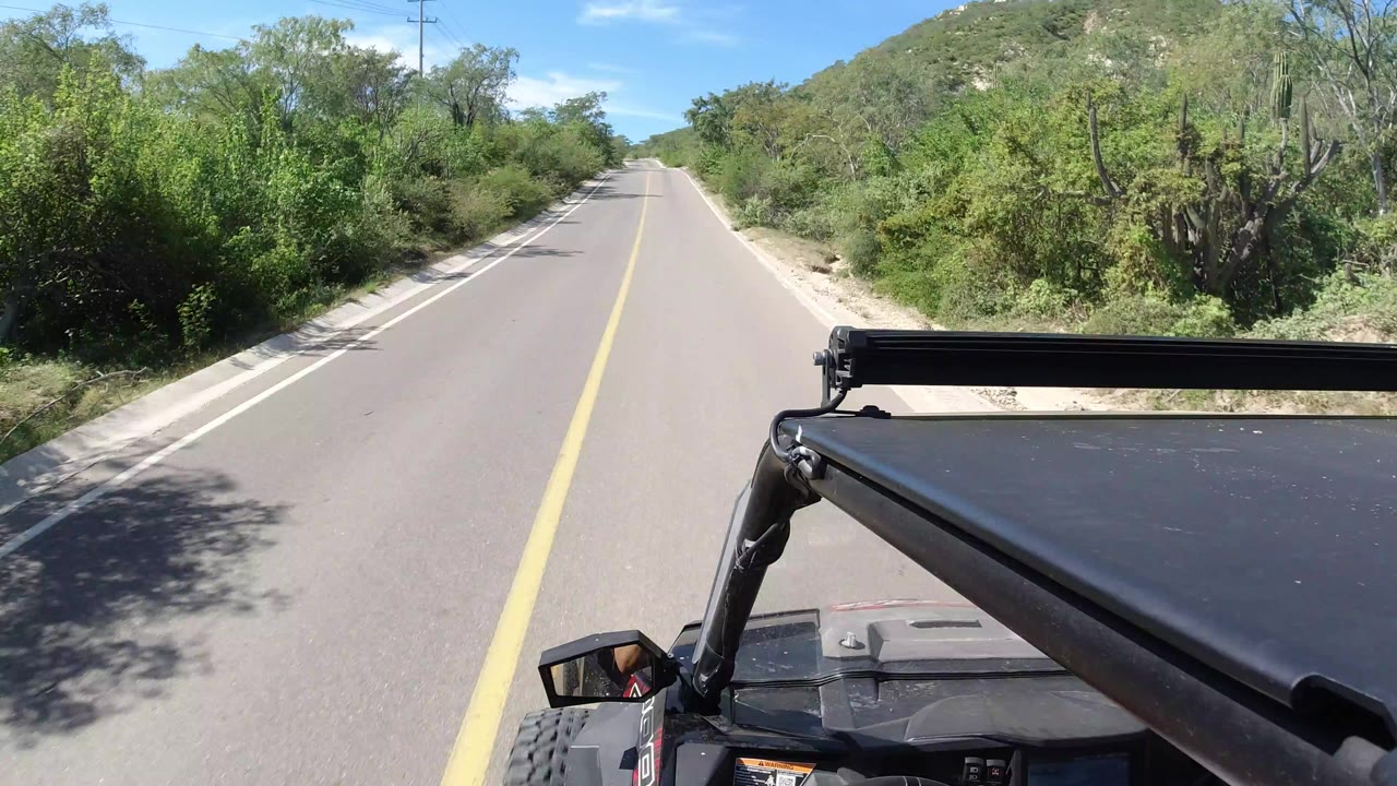 Forward view from an open vehicle on a Los Cabos road, with a yellow center line, green roadside brush, low hills, blue sky, and no visible people.