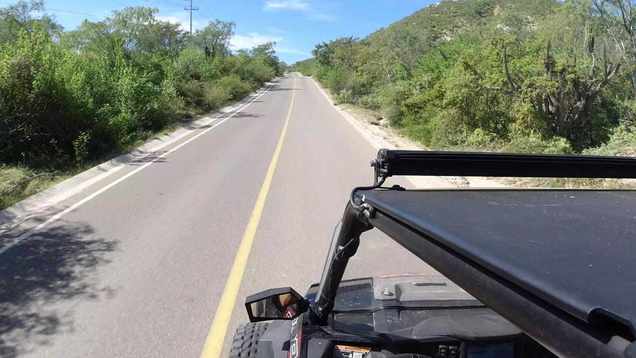The Los Cabos road continues ahead between thick green shrubs, with the vehicle's black roof support fixed across the lower right and no visible people.