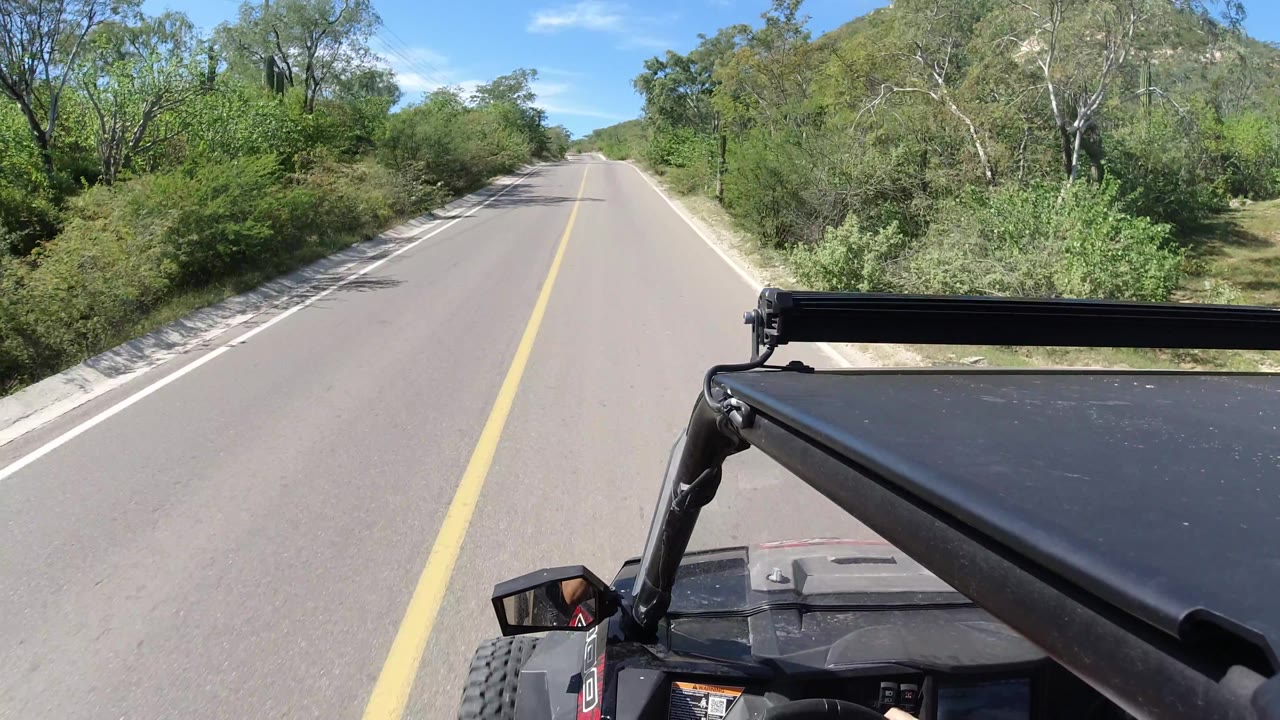 A forward-facing Los Cabos driving frame shows an empty road bending gently through brush, with hillside vegetation to the right and no visible people.