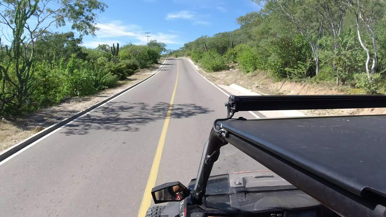 Forward road view in Los Cabos, Mexico, with a yellow center line, roadside trees, blue sky, and open vehicle hardware at lower right, with no visible people.