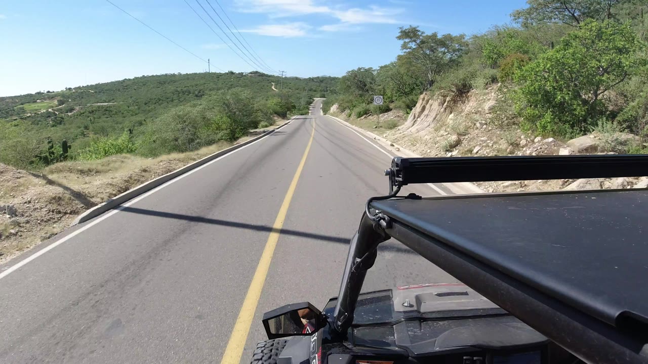 Second frame shows the same Los Cabos road running downhill through green scrub, with utility lines above, a rocky bank at right, the vehicle roof in the foreground, and no visible people.