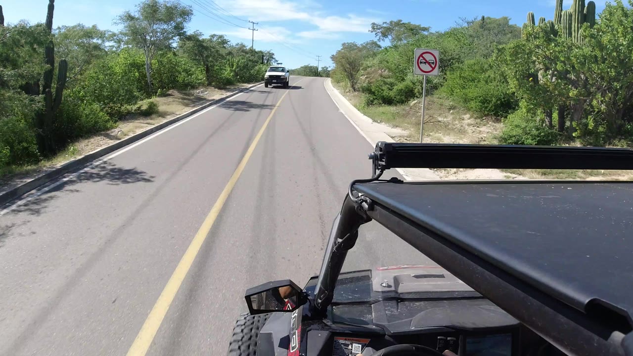Los Cabos road view from the Wrangler, showing the fixed roof bar in front, a no-symbol roadside sign, green vegetation, a distant vehicle, and no visible people.