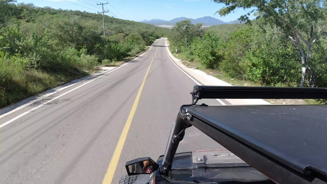 Forward vehicle view on a Los Cabos road with the Jeep roof and black bar in the foreground, green brush on both sides, distant mountains, and no visible people.