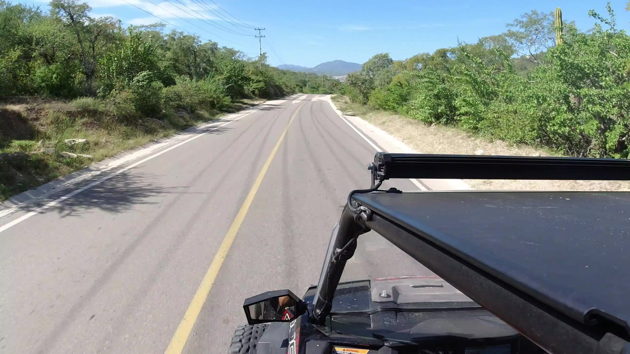 Forward-facing road view in Los Cabos, Mexico with a yellow centerline, green roadside brush, distant hills, part of a black vehicle in the foreground, and no visible people.