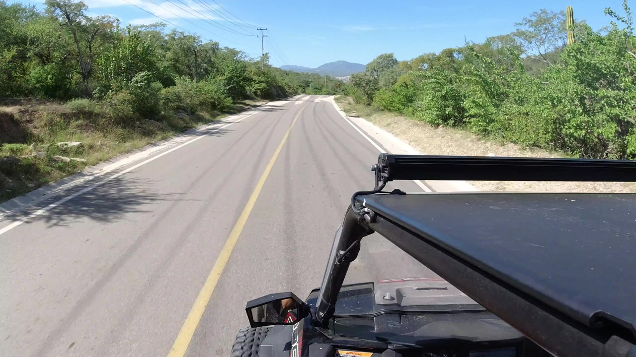 Los Cabos, Mexico road perspective with the black vehicle roof rack at lower right, shrubs close to the shoulder, low hills ahead, overhead utility lines, and no visible people.